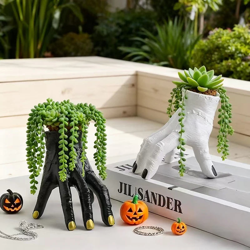 Two unique planters shaped like hands with green succulents on a surface with a book and Halloween-themed decorations.